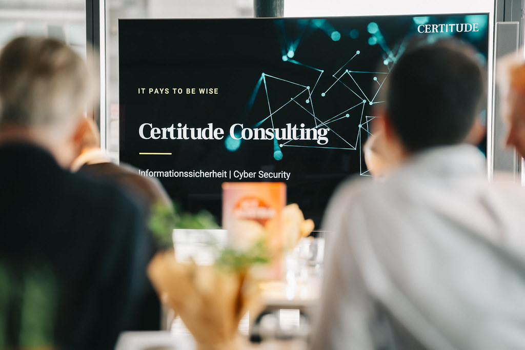 Presentation slide projected onto a screen, showing the text "Certitude Consulting" and "Pays to be wise", with blurred table decorations and attendees in the foreground.